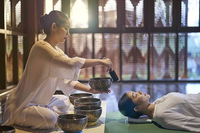 A therapist guiding guests through a sound bowl healing session in a sunlit garden courtyard.  